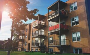 Brick apartment building with multiple balconies and a bicycle on the top balcony, sunlit lawn in the foreground.