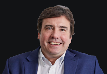 Head-and-shoulders portrait of a smiling man in a navy blazer and white shirt against a dark background.