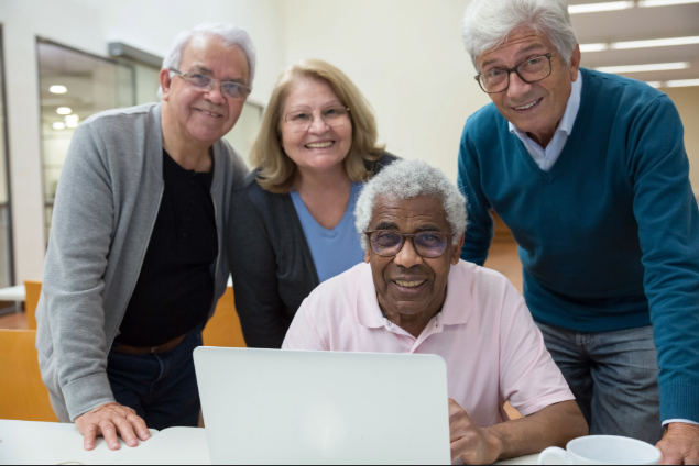 Four seniors gathered around a laptop, smiling at the camera in a bright room.