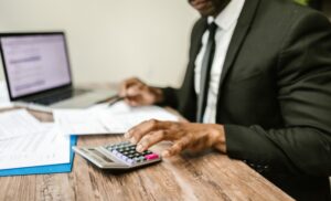Man in a dark suit using a calculator at a wooden desk with papers and a laptop nearby, focusing on finances.