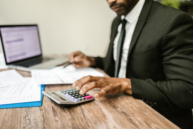 Man in a dark suit using a calculator at a wooden desk with papers and a laptop nearby, focusing on finances.