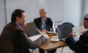 Business meeting around a wooden table with laptops, documents, and coffee cups, in a modern office setting.