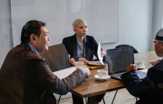 Business meeting around a wooden table with laptops, documents, and coffee cups, in a modern office setting.