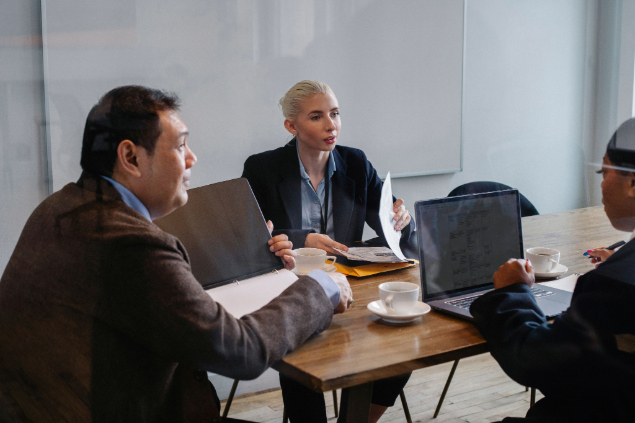 Business meeting around a wooden table with laptops, documents, and coffee cups, in a modern office setting.