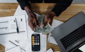 Person counting money with a calculator, laptop, and financial documents on a wooden desk.