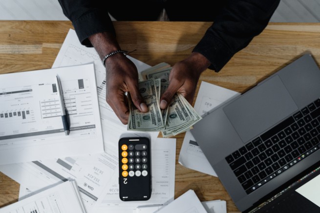 Person counting money with a calculator, laptop, and financial documents on a wooden desk.