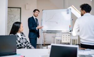 Man in a blue suit presents a line graph on a whiteboard to two attentive colleagues in a bright office space.