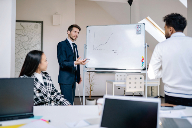 Man in a blue suit presents a line graph on a whiteboard to two attentive colleagues in a bright office space.