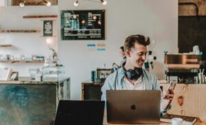 Man in a cafe coworking space, smiling at his phone with a laptop open in front of him and headphones around his neck.