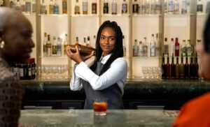 Female bartender in an apron shakes a cocktail shaker behind a bar, bottles lined on backlit shelves behind her.