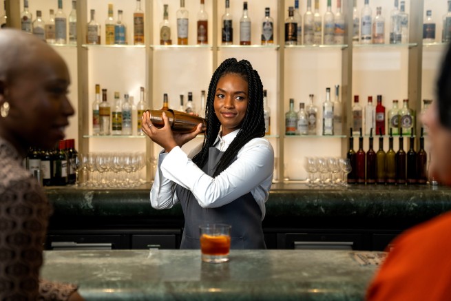Female bartender in an apron shakes a cocktail shaker behind a bar, bottles lined on backlit shelves behind her.