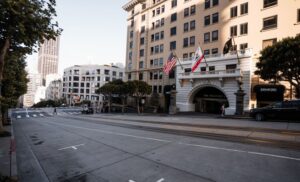 Facade of The Stanford Court hotel on a sunny San Francisco street, with flags at the entrance, parked cars, and tram tracks nearby.