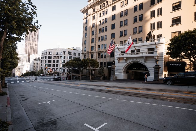 Facade of The Stanford Court hotel on a sunny San Francisco street, with flags at the entrance, parked cars, and tram tracks nearby.