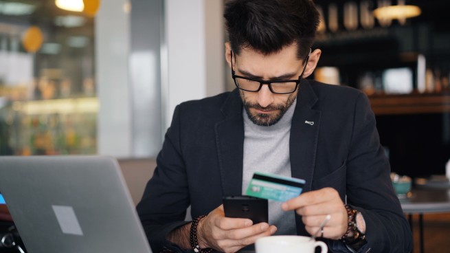 Man in a blazer and glasses pays online, using a credit card and smartphone beside an open laptop in a cafe-like setting.