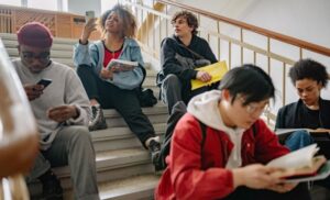 Diverse students sit on a school staircase, reading books, using a phone, or chatting during a study break.