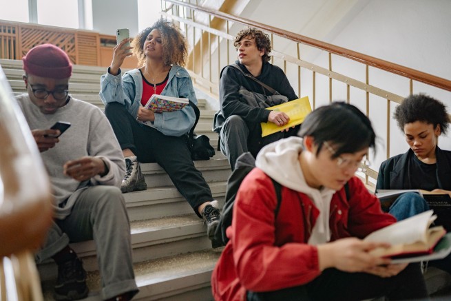 Diverse students sit on a school staircase, reading books, using a phone, or chatting during a study break.