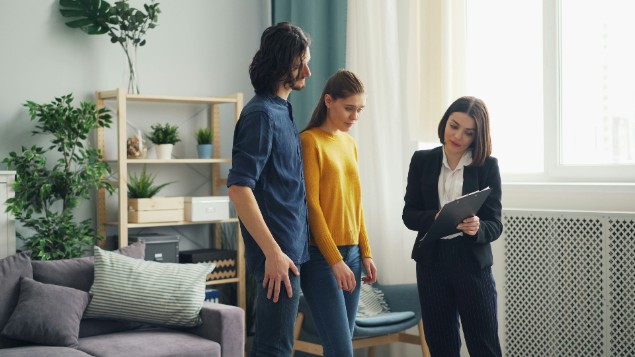 A woman in a blazer showing a clipboard to a couple in a bright living room, discussing documents.
