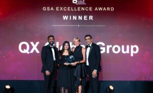 Group of four people in formal attire posing on stage with an award; backdrop reads 'GSA Excellence Award' and 'WINNER' with 'QX Group' visible in the background.