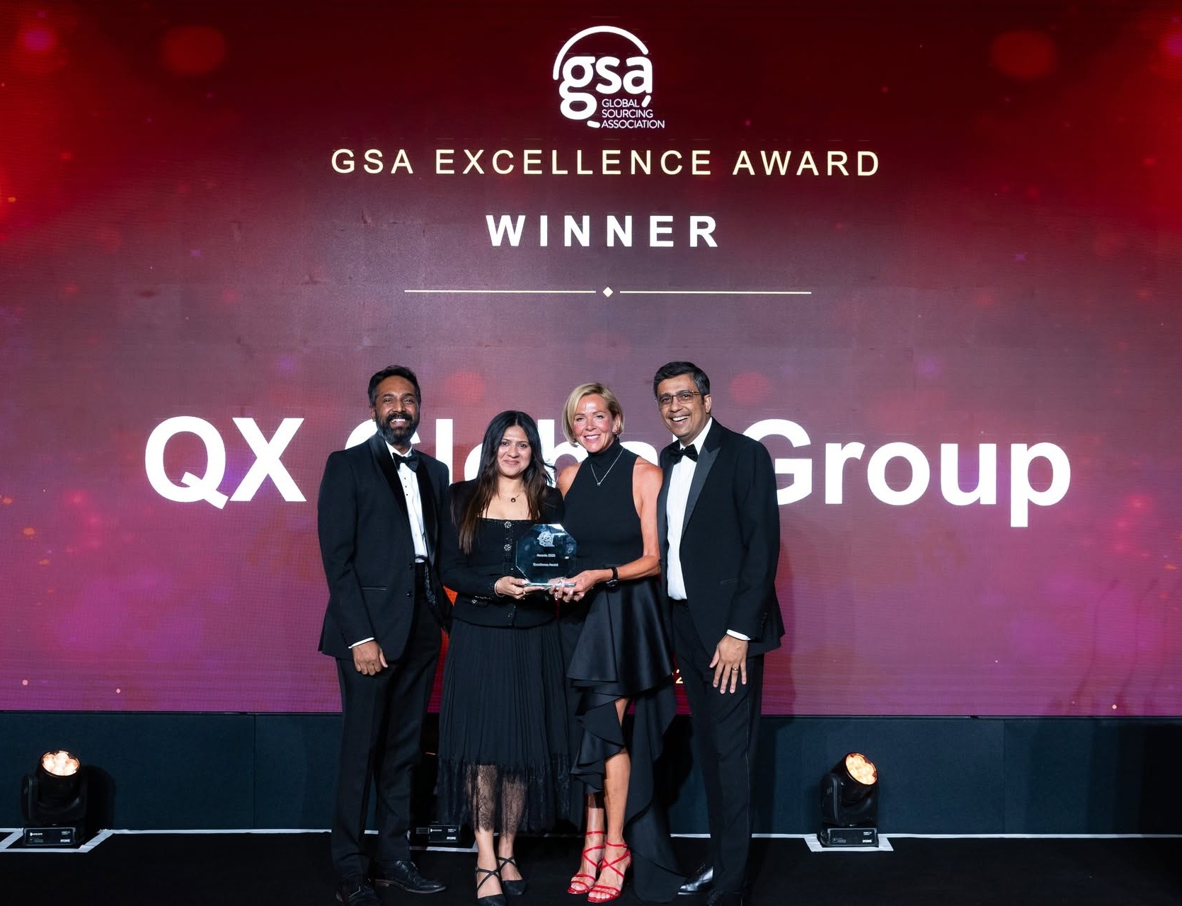 Group of four people in formal attire posing on stage with an award; backdrop reads 'GSA Excellence Award' and 'WINNER' with 'QX Group' visible in the background.