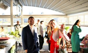Smiling man in a navy suit and a woman in a pink dress wave at the camera during a sunny rooftop party with guests mingling in the background.