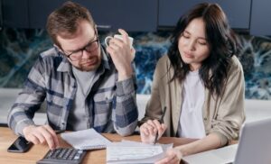 Two colleagues at a table reviewing documents; man with glasses and plaid shirt, woman writing notes, with a calculator, notebook, phone, and laptop nearby.